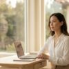 Composed professional woman in her 30s working calmly at a sunlit modern desk near a large window, polished exterior masking quiet inner pressure