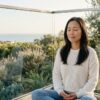 A calm woman sits cross-legged on a light-filled balcony overlooking a Southern California coastal view, eyes gently closed in a relaxed posture