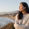 A person stands on a calm, empty beach at golden hour, looking out at gentle Pacific Ocean waves with warm light on the sand