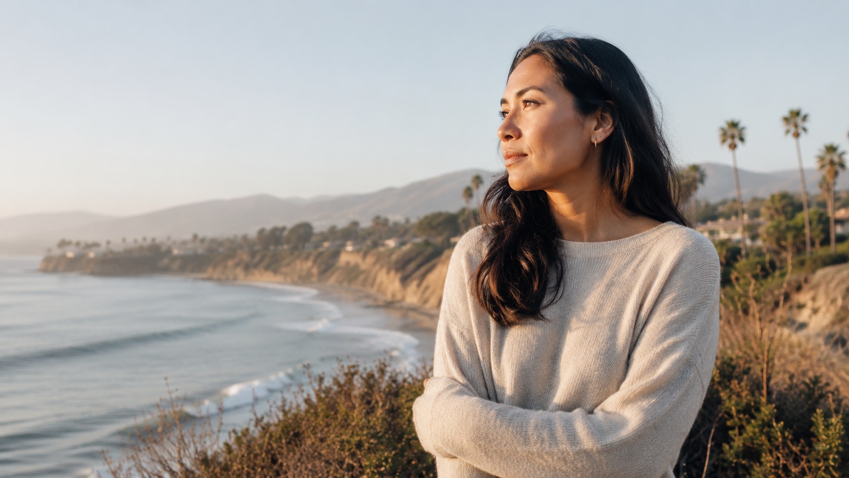 A person stands on a calm, empty beach at golden hour, looking out at gentle Pacific Ocean waves with warm light on the sand