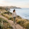 A person walks along a quiet coastal path in soft morning light, shoulders relaxed and unhurried