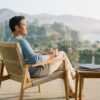 A professional sits quietly on a sunlit balcony with a coffee, looking out over soft coastal hills in calm morning light